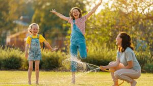 A mom spraying water and two happy girls jumping over it.