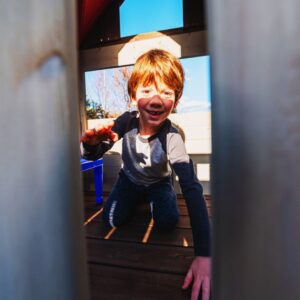 red haired kid smiles through slats in wooden playground 